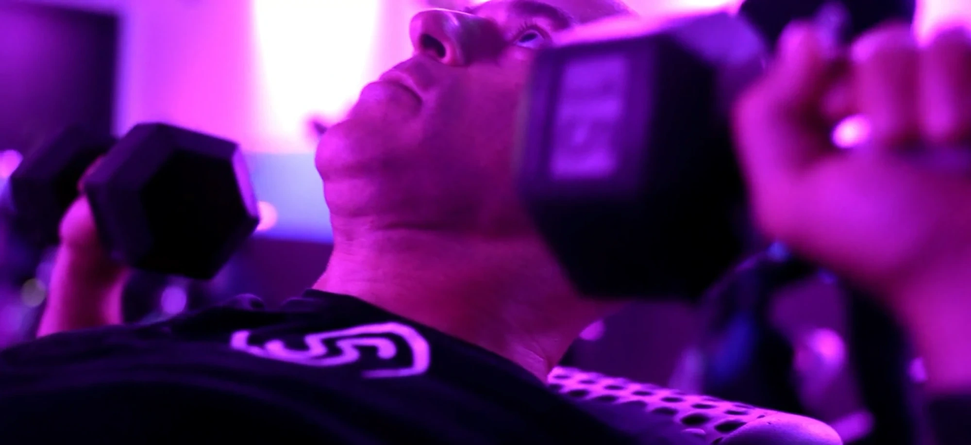 A Studio Spin member doing a dumbbell chest press on a bench under pink studio lights.