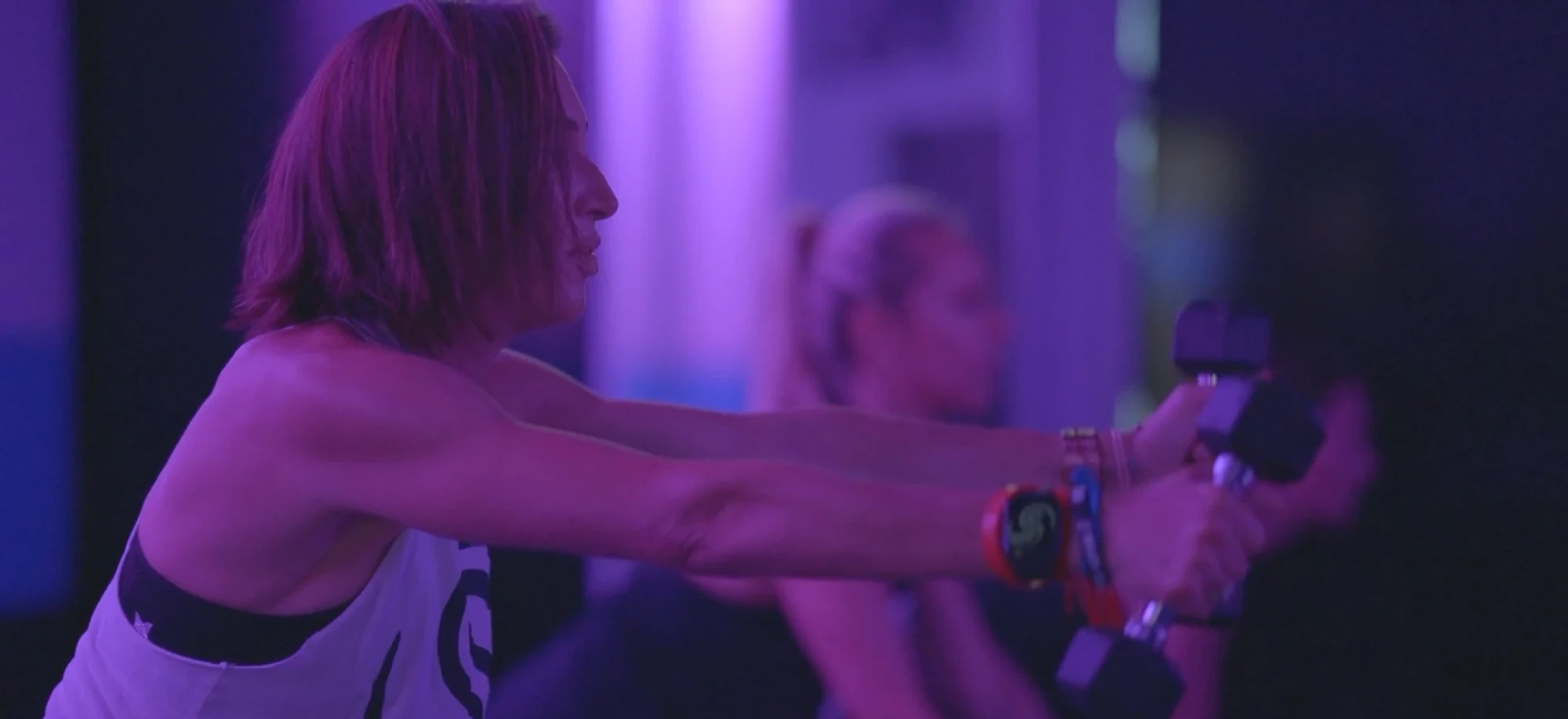 A Studio Spin member holding light dumbbells in a front raise under purple studio lights.