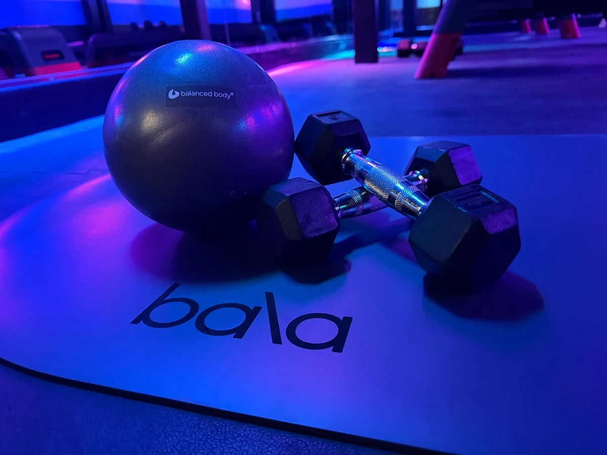 A Pilates ball and a pair of light dumbbells on an exercise mat under blue studio lights.
