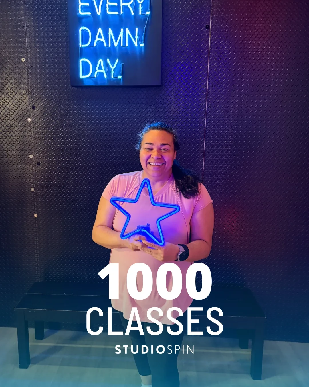 A Studio Spin member celebrating 1,000 classes under the Every Damn Day neon sign.