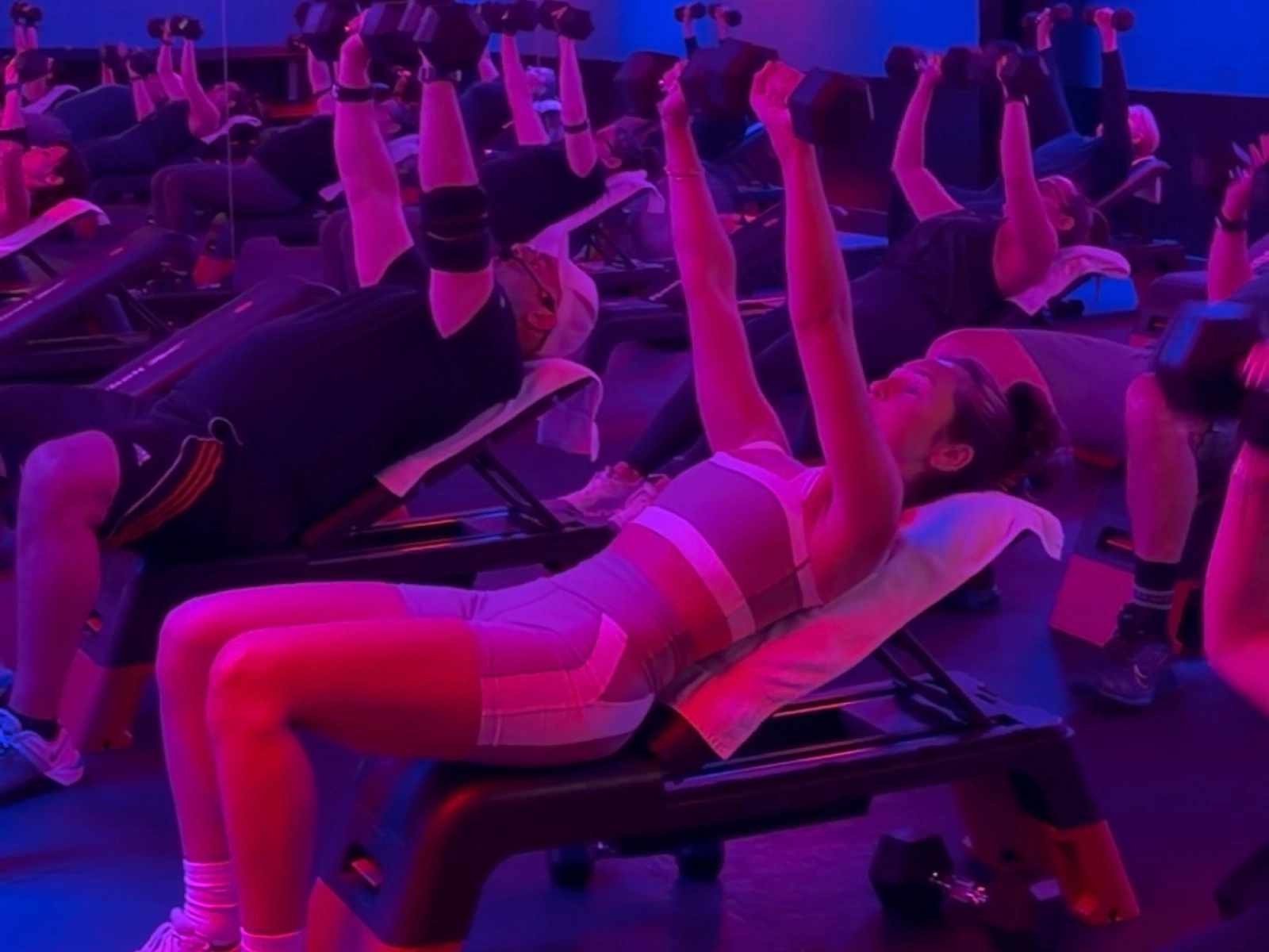 Studio Spin members performing dumbbell chest presses together under pink and blue studio lights.