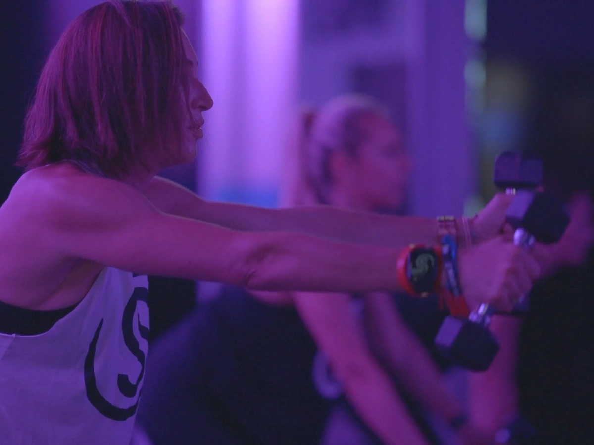 Studio Spin members performing dumbbell front raises together under purple studio lights.